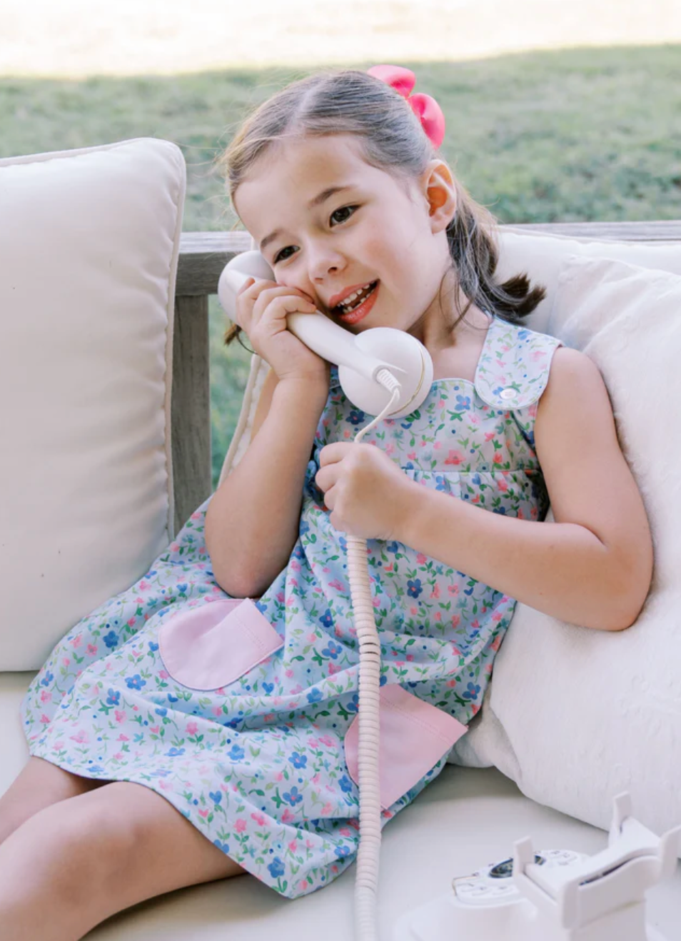Little girl in a blue and pink floral dress playfully pretending to have a phone conversation, smiling as she holds a toy phone to her ear.